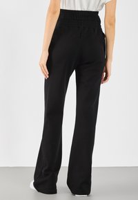 Schwarze, hochtaillierte Hose aus dehnbaren Stoff mit Schlagbeinen. Verfügt über einen elastischen Bund und seitliche Taschen.