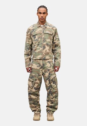 Uomo in piedi che indossa una giacca mimetica beige e verde e pantaloni cargo con scarpe beige con lacci, sfondo neutro.