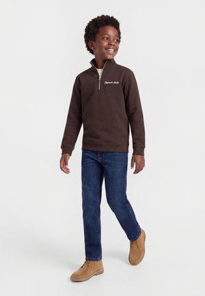 Jonge jongen loopt, draagt een bruin quarter-zip sweatshirt, blauwe spijkerbroek en beige laarzen, glimlacht en kijkt naar de zijkant.