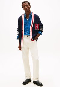 Marineblauer Herren-Strickjacke mit weißen und roten Streifen, versehen mit einem gestickten Logo. Getragen über einem blauen Jeanshemd und cremefarbenen Hosen mit schwarzen Stiefeln.