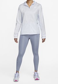 Persona vestida con una chaqueta con capucha de cremallera gris claro, leggings grises y zapatillas de correr blancas con detalles en rosa, de pie contra un fondo blanco.