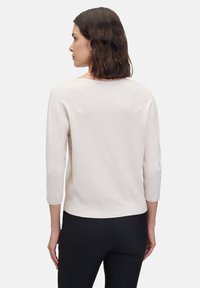 Hellbeige gestreifter Pullover mit dreiviertelÄrmeln und einem lockeren Schnitt, ausgestattet mit einem weiten Ausschnitt und subtilem Struktur. Schwarze Hose ist sichtbar.