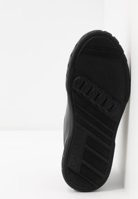 Semelle de sneaker en caoutchouc noir avec des motifs texturés et des rainures de flexion, comportant une empreinte de logo sur le côté et une zone de talon lisse et plate.