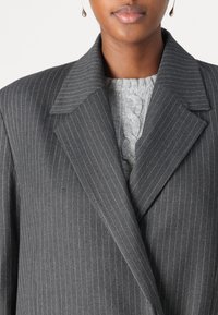 Cappotto grigio a righe con colletto ampio, con un morbido maglione grigio a treccia sotto. Vista ravvicinata della parte superiore degli indumenti.