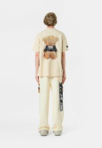 Camiseta de algodón beige con un gráfico de osito de peluche texturizado en la parte posterior. Combinada con pantalones color crema y detalles de tela únicos.