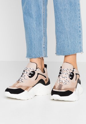 Chunky sneakers in beige, black, and white with patterned laces worn with frayed hem light blue denim jeans.