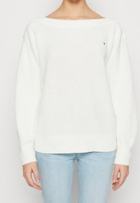 Maglione bianco lavorato a maglia con scollatura a spalle scoperte, maniche ampie e polsini a coste. Presenta un piccolo logo sul petto.