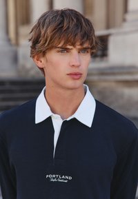 Tumesinise värviga polo-särk valge kraega, millel on tikitud "PORTLAND" logo. Sooniline tekstuur klassikalise lõike ja kolme nööpauguga.