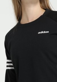 Chemise noire à manches longues avec un logo Adidas blanc sur la poitrine et trois bandes blanches sur la manche gauche. Tissu doux, coupe décontractée.