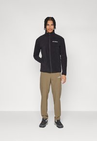 Svart fleece-zip-jakke med høy krage; paret med olivengrønne bukser med sidelommer og merkede detaljer. Svart fottøy.