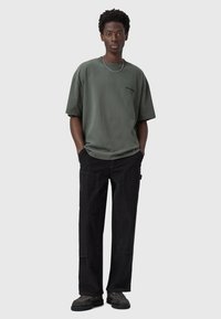 Camiseta de algodón verde oliva de gran tamaño con un logo negro, combinada con pantalones de pana negros que cuentan con bolsillos, un acabado texturizado y piernas anchas.