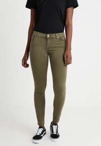 Mujer vestida con jeans ajustados en color verde oliva, camiseta negra y zapatillas en blanco y negro, de pie contra un fondo sencillo.
