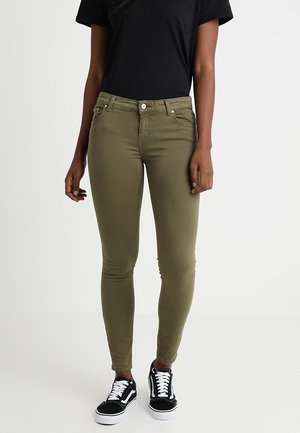 Mujer vestida con jeans ajustados en color verde oliva, camiseta negra y zapatillas en blanco y negro, de pie contra un fondo sencillo.