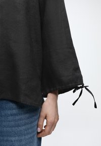 Χέρι με ανοιχτό μωβ βερνίκι νυχιών, μερικώς καλυμμένο από μαύρο φαρδύ μακρυμάνικο τοπ με δεμένο κορδόνι στο μανίκι, συνδυασμένο με μπλε τζιν παντελόνι.