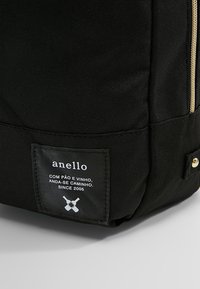 Zaino in tessuto nero con una targhetta frontale ben visibile, cerniera dorata e materiale testurizzato senza motivi o decorazioni visibili.