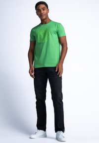 Camiseta de algodón verde con un estampado gráfico circular en el frente, combinada con pantalones de mezclilla negros y zapatillas blancas. Estilo simple y casual.