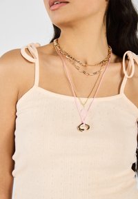 Beige ribbat topp med axelband, prydd med flera lager av halsband i guld och rosa med varierande texturer och mönster.