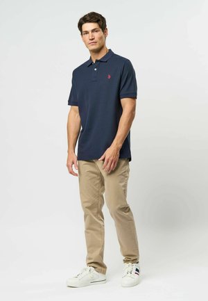 Tumesinine polo särk lühikeste krae ja kolme nööbiga, koos beežide chinos-pükstest ja valgetest tossudest, millel on sinised ja punased aktsendid.