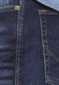 Țesătură de denim de culoare albastru închis, cu un buzunar frontal vizibil, cu cusături galbene în contrast și un detaliu de buton metalic. Textură netedă cu o ușoară decolorare.