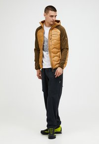 Padded jacket in tan and green, featuring a zip front, hood, and a quilted design. Paired with dark pants and hiking boots.