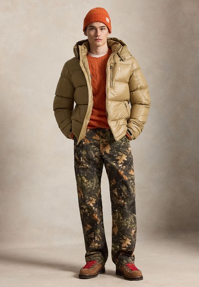 Jeune homme portant un bonnet en maille orange, une doudoune beige, un pull orange, un pantalon camouflage et des chaussures de randonnée marron avec des lacets rouges.