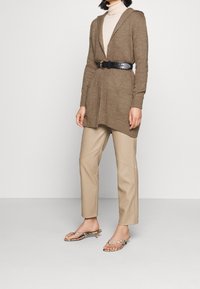 Hnědý pletený svetr s kapucí, se zavazováním v pase. Béžové upravené kalhoty s detaily švů. Průhledné sandály na podpatku se pásky.