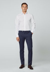 Weißes Langarmhemd mit Knopfverschluss, kombiniert mit dunkelblauen, taillierten Hosen mit einem dezenten Karomuster. Braune Lederschuhe sind ebenfalls enthalten.