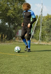 Schwarzer Sportanzug mit blauen und grünen Akzenten; Spieler dribbelt einen mehrfarbigen Fußball auf einem Rasenfeld.