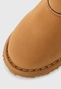 Botim de tornozelo em camurça bege, com ponta arredondada, apresentando uma textura suave, costura contrastante e uma sola de borracha durável.