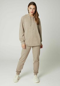 Felpa beige con cappuccio e cordino, maniche lunghe e vestibilità rilassata; abbinata a pantaloni affusolati coordinati. Indossata con sneakers sportive bianche.