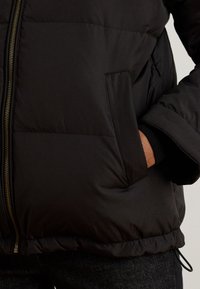 Μαύρο μπουφάν τύπου puffer με φερμουάρ, πλαϊνές τσέπες και ριμπ μανσέτες. Λείο, ελαφρύ ύφασμα με καπιτονέ υφή.