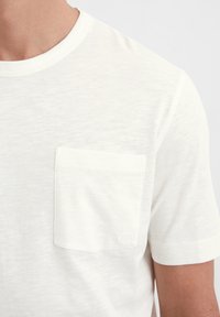 Weißes T-Shirt mit kurzen Ärmeln aus leichtem Stoff. Mit einer Brusttasche und einem dezenten Logo sowie einem runden Ausschnitt. Weicher Stoff.