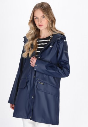 Frau mit langen, gewellten blonden Haaren, die einen marineblauen Regenmantel über einem schwarz-weiß gestreiften Oberteil trägt und vor einem weißen Hintergrund steht.