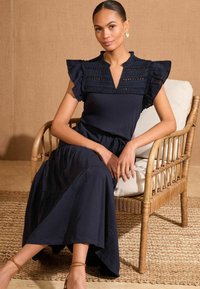 Rochie bleumarin cu detalii din dantelă la decolteu, mâneci volănaș și fustă fluidă. Purta sandale cu curele, așezată pe un scaun din rachită.