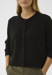 Frau in einer schwarzen, knöpfbaren Strickjacke mit langen, bis zu den Ellbogen gerollten Ärmeln und olivgrünen Hosen, Hände in den Taschen, vor einem einfarbigen Hintergrund.