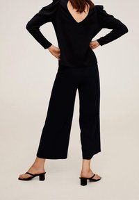 Černý dlouhý rukáv top s hlubokým V-výstřihem na zádech, spárovaný s širokými kalhotami. Materiál je hladký a lehký; boty jsou ploché sandály s blokovým podpatkem.