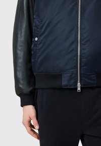 Navy bomber jacket with a sleek satin body and black leather sleeves. Features a silver zipper and ribbed cuffs, with a side pocket.