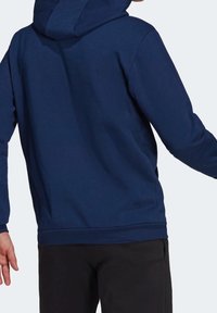 Sweat à capuche bleu marine avec cordon de serrage, ourlet et poignets côtelés, et une texture douce. Porté avec un pantalon noir, affichant une coupe décontractée.