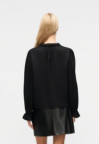 Γυναίκα με μακριά ξανθά μαλλιά που φοράει μαύρο πουκάμισο με μακριά μανίκια και βολάν στις μανσέτες και μαύρη δερμάτινη φούστα, εμφανιζόμενη από πίσω.