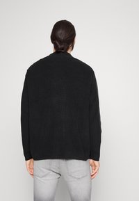 Schwarzer gerippter Strickpullover mit lockerer Passform, Rundhalsausschnitt und langen Ärmeln, von hinten betrachtet. Kombiniert mit grauen Jeans.