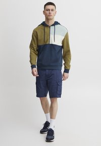 Felpa con cappuccio in blocchi di colore oliva, azzurro e blu navy, con tasca frontale, abbinata a pantaloni cargo blu navy e scarpe da atletica nere.