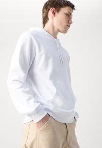 Felpa bianca realizzata in tessuto morbido, con tasca a marsupio frontale, cappuccio regolabile con cordino e un piccolo logo dorato sul petto.