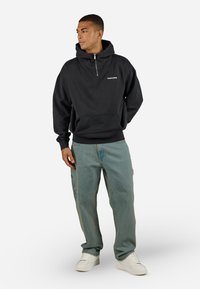Zwarte pullover hoodie met een half-rits halslijn, een kangoeroezak en een wit logo; gecombineerd met licht blauwe baggy jeans en witte sneakers.