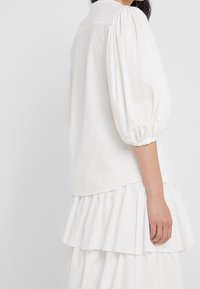 Blusa bianca con maniche a sbuffo e orlo arricciato a strati. Tessuto testurizzato con sottili righe, dotato di un colletto classico.