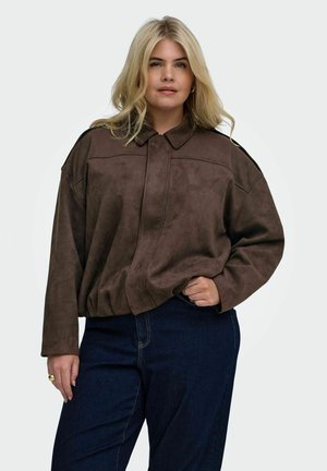 Blonde Frau, die eine lockere braune Jacke und dunkelblaue Jeans trägt, steht mit einer Hand in der Tasche vor einem einfarbigen Hintergrund.