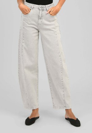 Personne portant un jean gris clair à jambes larges et des chaussures plates noires pointues, mains dans les poches avant, debout sur un fond gris.