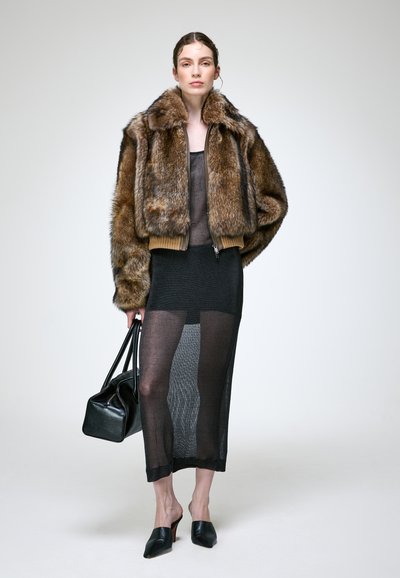 Femme portant une veste en fourrure marron, une robe noire transparente, des talons noirs, tenant un sac à main noir, debout devant un fond clair uni.
