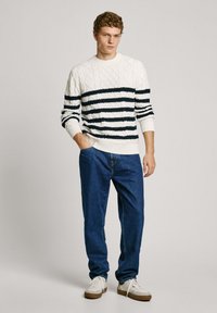 Pull en maille blanche à rayures navy, avec une texture en cable et des poignets côtelés. Associé à un jean en denim bleu et des baskets blanches.