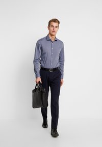 Męska jasnoniebieska koszula z długim rękawem, granatowe spodnie, czarny pasek i czarna skórzana torba, idąca na neutralnym tle.