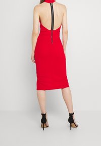 Röd kroppsnära klänning med hög krage, öppen rygg och en svart dragkedjedetalj. Midjan når till mitten av vaderna och kombineras med svarta högklackade skor.